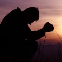  Hay una persona en silueta arrodillada con las manos entrelazadas en oración al atardecer, transmitiendo fe, rendición, esperanza y búsqueda de fortaleza en medio de la lucha contra la adicción.