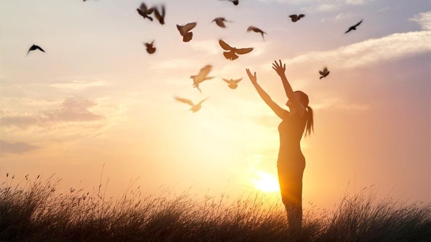 Niña al atardecer con los brazos levantados mientras suelta las aves al cielo, simbolizando libertad, soltar el control, esperanza y paz interior.
